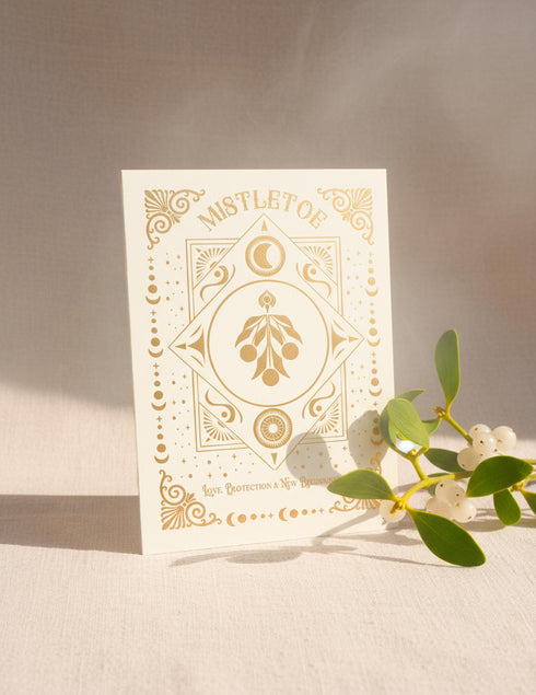 A Grimoire & Goblet luxury Mistletoe ritual card set. The 5x7 cream greeting card features gold celestial designs framing “Mistletoe” and “Love, Protection & New Beginnings.” Shown with a vellum wrap printed in gold florals, a white wax seal with mistletoe, and a premium envelope. Includes a 4x6 ritual insert on handmade deckled paper titled “Ritual for Peace and Protection.” A spiritual Yule stationery set designed as a luxurious unboxing experience.
