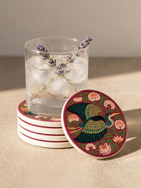 A round, 4-inch ceramic drink coaster featuring a glossy, flat-printed Art Nouveau design. The central motif is a flying heron with teal plumage and scalloped golden wing feathers, surrounded by stylized winding olive green foliage, peach chrysanthemums, and botanical blooms. The artwork is set against a solid, deep blood-red background. The coaster has a smooth white edge and a natural brown cork backing. Available as a single coaster or in matching sets of four or six.