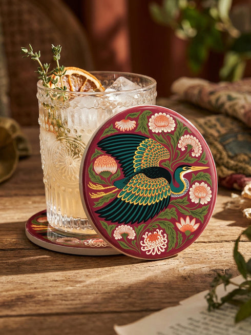 A round, 4-inch ceramic drink coaster featuring a glossy, flat-printed Art Nouveau design. The central motif is a flying heron with teal plumage and scalloped golden wing feathers, surrounded by stylized winding olive green foliage, peach chrysanthemums, and botanical blooms. The artwork is set against a solid, deep blood-red background. The coaster has a smooth white edge and a natural brown cork backing. Available as a single coaster or in matching sets of four or six.
