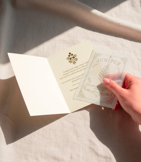a luxury Grimoire and Goblet ritual card set. A 5x7 cream-colored folded card features an intricate, art-nouveau-style gold foil illustration of a Juniper plant, with the words "Juniper: Cleansing & Fortune." The card is presented with a translucent vellum envelope, which has a gold floral border and a white adhesive wax seal. Inside, the card reveals a vellum-wrapped keepsake, which unfurls to a 4x6 ritual card printed on thick, 160 GSM handmade cotton paper with a rough, deckled edge.
