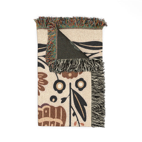 Grimoire and Goblet woven jacquard cotton blanket / wall tapestry featuring a centered folk-art moth framed by symmetrical wildflowers and leafy stems. Natural oatmeal-beige base with rust/terracotta and cocoa-brown moth detail, charcoal-black botanicals, and warm ivory highlights. Finished with a 2-inch multicolor fringe and a reversible color-negative back. Available in 52x37, 50x60, and 60x80 inches as a throw, bed cover, or statement wall hanging.