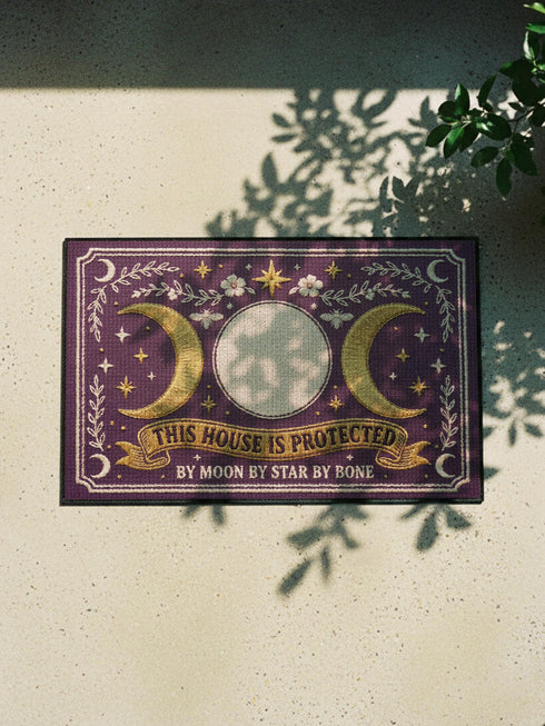 A rectangular doormat featuring a printed faux embroidery design on a deep plum purple background. The symmetrical artwork centers on a large cream full moon flanked by two golden crescent moons, creating a Triple Moon symbol. Above the moons are gold stars, cream flowers, and two small bees. Below, a golden scroll banner reads "THIS HOUSE IS PROTECTED" with "BY MOON BY STAR BY BONE" underneath. The mat is framed by cream leafy vines and finished with a black fabric border trim.