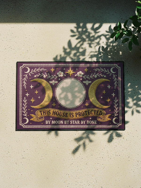 A rectangular doormat featuring a printed faux embroidery design on a deep plum purple background. The symmetrical artwork centers on a large cream full moon flanked by two golden crescent moons, creating a Triple Moon symbol. Above the moons are gold stars, cream flowers, and two small bees. Below, a golden scroll banner reads "THIS HOUSE IS PROTECTED" with "BY MOON BY STAR BY BONE" underneath. The mat is framed by cream leafy vines and finished with a black fabric border trim.