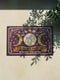 A rectangular doormat featuring a printed faux embroidery design on a deep plum purple background. The symmetrical artwork centers on a large cream full moon flanked by two golden crescent moons, creating a Triple Moon symbol. Above the moons are gold stars, cream flowers, and two small bees. Below, a golden scroll banner reads "THIS HOUSE IS PROTECTED" with "BY MOON BY STAR BY BONE" underneath. The mat is framed by cream leafy vines and finished with a black fabric border trim.