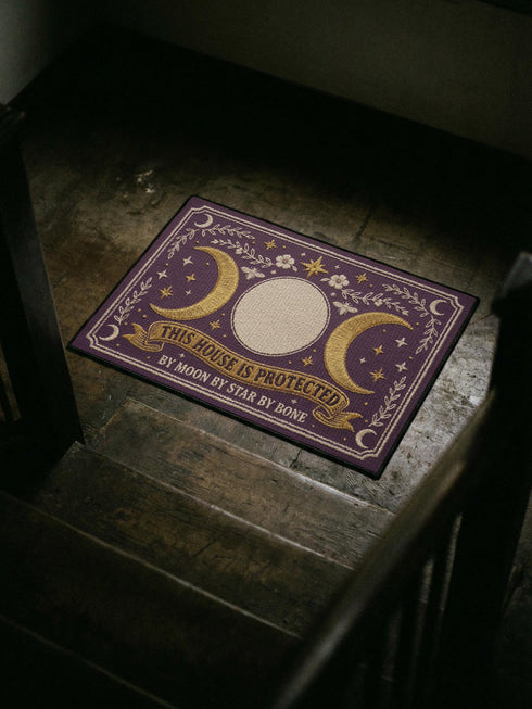 A rectangular doormat featuring a printed faux embroidery design on a deep plum purple background. The symmetrical artwork centers on a large cream full moon flanked by two golden crescent moons, creating a Triple Moon symbol. Above the moons are gold stars, cream flowers, and two small bees. Below, a golden scroll banner reads "THIS HOUSE IS PROTECTED" with "BY MOON BY STAR BY BONE" underneath. The mat is framed by cream leafy vines and finished with a black fabric border trim.
