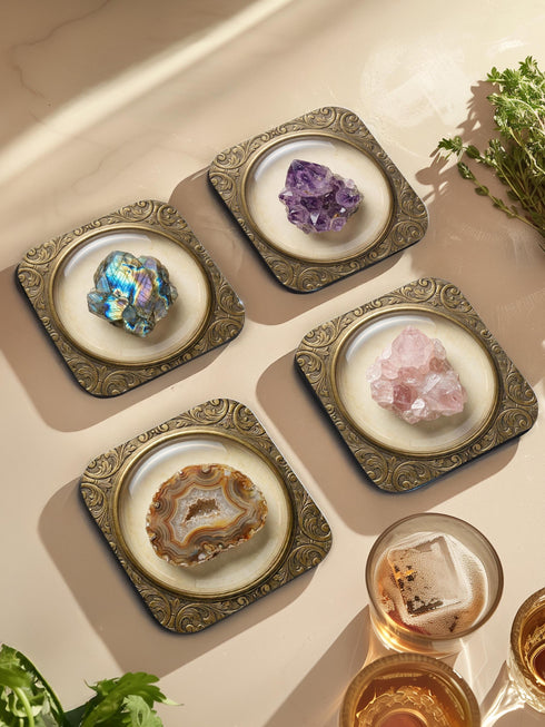 Grimoire and Goblet set of four square glossy coasters featuring high-resolution printed illustrations of raw crystals: purple amethyst, pink rose quartz, iridescent labradorite, and banded agate. Each gemstone is depicted inside a printed faux antique brass baroque frame with intricate scrollwork, set against a cream parchment background. The design is a flat 2D trompe l'œil print on hardboard, creating a realistic 3D specimen box illusion.