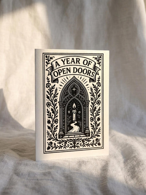 A vertical 5x7 inch pearlescent greeting card by Grimoire and Goblet featuring a high-contrast black woodcut-style illustration. The design centers on a gothic cathedral archway framing a glowing candle and a winding path leading forward. Surrounding the arch are intricate floral vines and stars. A banner at the top reads A YEAR OF OPEN DOORS in serif typography. The paper has a subtle silvery shimmer. The inside contains a printed poem about warmth and promise. Perfect for spiritual gifting.