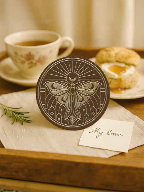 A round, dark brown ceramic drink coaster with a non-slip cork back. The top surface features an intricate, engraved-style celestial moth design in a light beige or off-white color. The luna moth is centered under a crescent moon and a radiant sunburst. The design is symmetrical and includes stars and geometric line art, evoking a gothic, witchy, and spiritual aesthetic. The coaster has a matte finish and is designed as a piece of functional art.