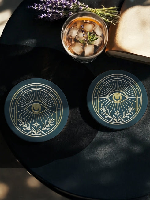 A round ceramic coaster with a smooth, matte, dark slate-blue surface and a natural, textured cork backing. The top of the coaster features an intricate, metallic gold line-art design of the All-Seeing Eye, also known as the Eye of Providence. A central eye with a defined iris and pupil is enclosed within a circular emblem, from which fine golden lines radiate outwards like beams of light. Below the eye, two stylized laurel branches curve upwards, cradling a small, four-pointed star.