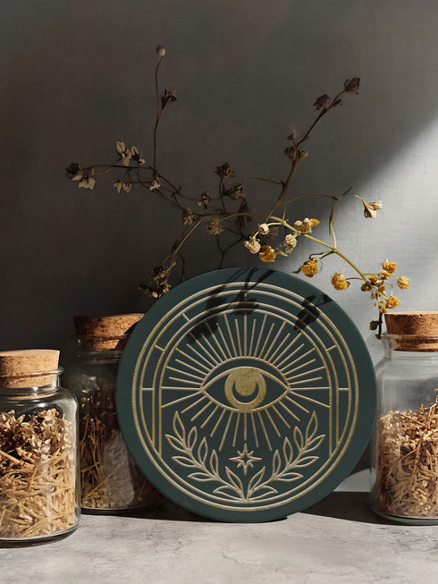 A round ceramic coaster with a smooth, matte, dark slate-blue surface and a natural, textured cork backing. The top of the coaster features an intricate, metallic gold line-art design of the All-Seeing Eye, also known as the Eye of Providence. A central eye with a defined iris and pupil is enclosed within a circular emblem, from which fine golden lines radiate outwards like beams of light. Below the eye, two stylized laurel branches curve upwards, cradling a small, four-pointed star.