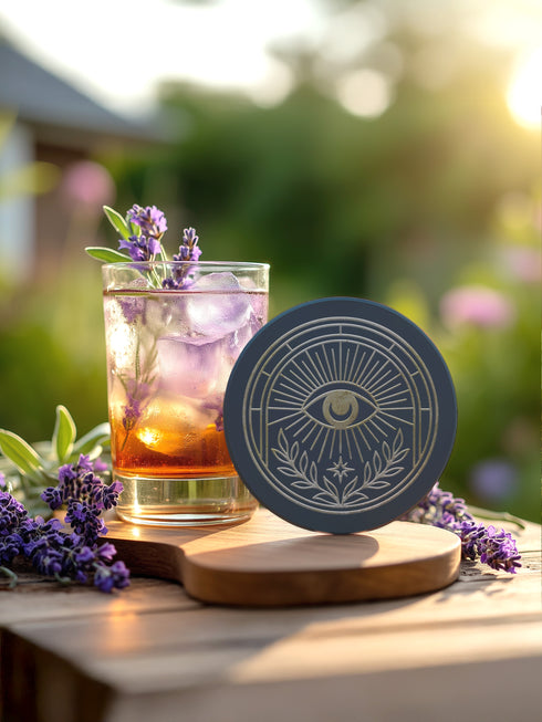 A round ceramic coaster with a smooth, matte, dark slate-blue surface and a natural, textured cork backing. The top of the coaster features an intricate, metallic gold line-art design of the All-Seeing Eye, also known as the Eye of Providence. A central eye with a defined iris and pupil is enclosed within a circular emblem, from which fine golden lines radiate outwards like beams of light. Below the eye, two stylized laurel branches curve upwards, cradling a small, four-pointed star.