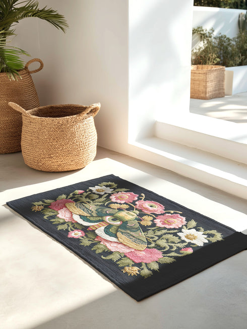 Dark charcoal doormat featuring an ornate death moth with spread wings at center, surrounded by intricate botanical border of pink roses, white daisies, and golden wheat stalks in symmetrical folk art design on textured woven chenille surface. The moth displays detailed wing patterns with eye-like markings and striped body, positioned as central focal point within lush floral frame that includes delicate leaves in sage green and teal tones.