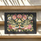 Dark charcoal doormat featuring an ornate death moth with spread wings at center, surrounded by intricate botanical border of pink roses, white daisies, and golden wheat stalks in symmetrical folk art design on textured woven chenille surface. The moth displays detailed wing patterns with eye-like markings and striped body, positioned as central focal point within lush floral frame that includes delicate leaves in sage green and teal tones.