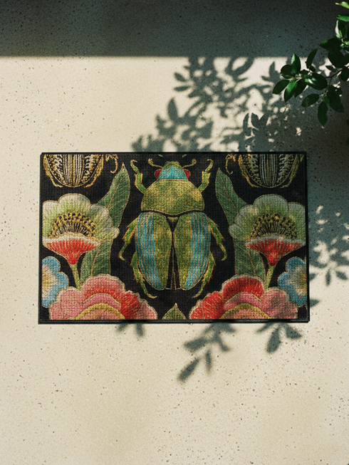 A rectangular doormat featuring a high-fidelity faux embroidery print on a solid black background. The central design is a large, detailed green and teal scarab beetle. It is surrounded by vibrant, flat-printed botanical motifs including large pink, red, and light blue flowers, green foliage, and pale gold-toned seed pods. The mat features a black fabric trim around the edges. The colorful print creates a 2D trompe-l'œil illusion of raised tapestry needlework on a smooth polyester surface.