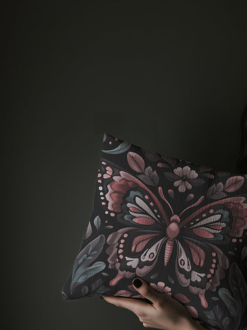 A square decorative pillow featuring an intricately detailed butterfly design in rose pink, blush, and silver-grey colors against a black background. The butterfly is surrounded by botanical elements with symmetrical floral patterns.