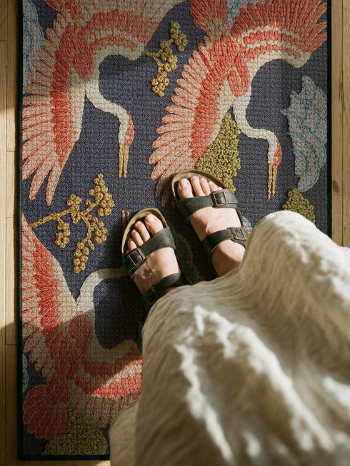 Grimoire and Goblet chinoiserie flying crane faux embroidery doormat featuring three cranes in coral blush and jade green soaring across deep navy background with teal scalloped clouds golden bonsai branches and olive foliage. Printed polyester entryway mat with non-slip rubber backing and black trim. Available 24x18 and 36x24 inches. Asian inspired Japanese crane design housewarming gift home decor