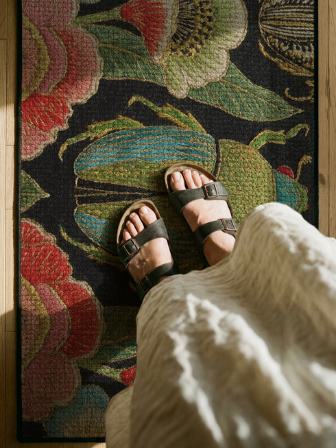 A rectangular doormat featuring a high-fidelity faux embroidery print on a solid black background. The central design is a large, detailed green and teal scarab beetle. It is surrounded by vibrant, flat-printed botanical motifs including large pink, red, and light blue flowers, green foliage, and pale gold-toned seed pods. The mat features a black fabric trim around the edges. The colorful print creates a 2D trompe-l'œil illusion of raised tapestry needlework on a smooth polyester surface.