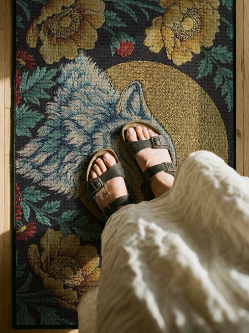 Grimoire and Goblet rectangular doormat featuring a faux embroidery style printed design of a howling wolf with blue and ivory fur, centered in front of a textured antique gold circular moon disc, surrounded by large golden yellow flowers, teal leaves, and small red berry accents on a dark charcoal background. Entryway rug with black trim, non slip backing, and botanical nature inspired mystical design.