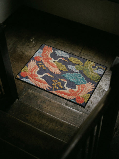 Grimoire and Goblet chinoiserie flying crane faux embroidery doormat featuring three cranes in coral blush and jade green soaring across deep navy background with teal scalloped clouds golden bonsai branches and olive foliage. Printed polyester entryway mat with non-slip rubber backing and black trim. Available 24x18 and 36x24 inches. Asian inspired Japanese crane design housewarming gift home decor