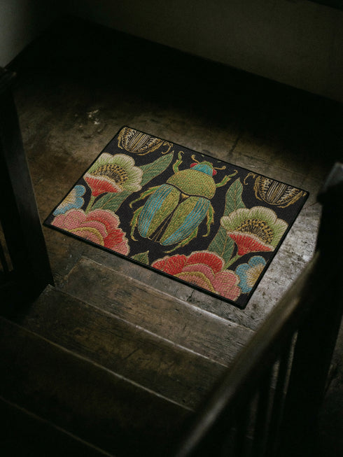 A rectangular doormat featuring a high-fidelity faux embroidery print on a solid black background. The central design is a large, detailed green and teal scarab beetle. It is surrounded by vibrant, flat-printed botanical motifs including large pink, red, and light blue flowers, green foliage, and pale gold-toned seed pods. The mat features a black fabric trim around the edges. The colorful print creates a 2D trompe-l'œil illusion of raised tapestry needlework on a smooth polyester surface.
