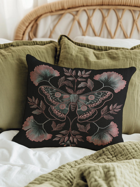 A square decorative pillow featuring an intricately detailed Luna moth in faded rose pink, slate blue and dusty terracotta colors on a black background. The moth is centered in a symmetrical arrangement with ginkgo-inspired leaves and botanical elements surrounding it.