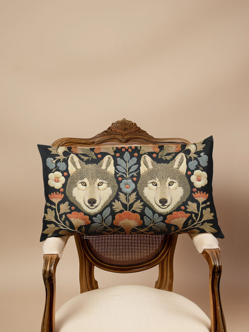 A rectangular black lumbar pillow, 12x20 inches, with a detailed symmetrical folk art print. The design features two forward-facing grey wolf heads in the center, flanked by intricate botanical elements. The color palette is moody and rustic, with flowers in burnt orange and cream, and leaves in slate blue and antique gold. The artwork has a faux embroidery, tapestry-style texture. This woodland animal accent cushion is made from a premium polyester fabric with a soft, linen feel.
