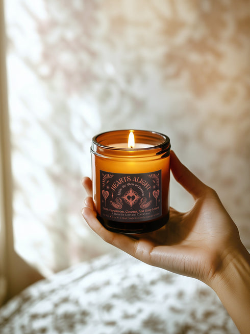 A 9 oz amber glass candle featuring a black label with rose gold art depicting hearts and swans. The label reads "Hearts Alight" and lists cardamom, coconut, and lemon fragrance notes. A cotton wick burns at the center.