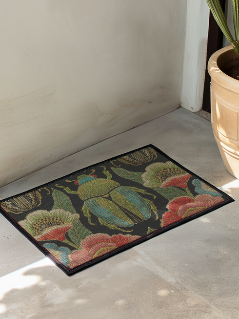 A rectangular doormat featuring a high-fidelity faux embroidery print on a solid black background. The central design is a large, detailed green and teal scarab beetle. It is surrounded by vibrant, flat-printed botanical motifs including large pink, red, and light blue flowers, green foliage, and pale gold-toned seed pods. The mat features a black fabric trim around the edges. The colorful print creates a 2D trompe-l'œil illusion of raised tapestry needlework on a smooth polyester surface.