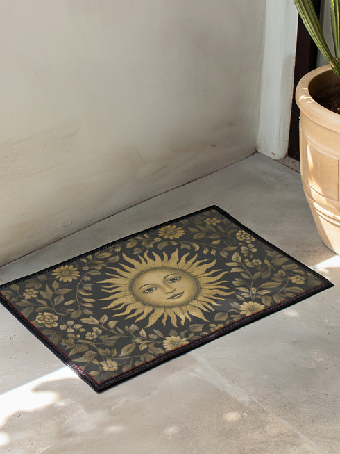 Handcrafted celestial doormat featuring an antique gold sun face with knowing eyes, surrounded by botanical motifs against a deep charcoal background with elegant black trim.
