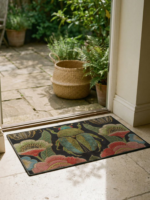 A rectangular doormat featuring a high-fidelity faux embroidery print on a solid black background. The central design is a large, detailed green and teal scarab beetle. It is surrounded by vibrant, flat-printed botanical motifs including large pink, red, and light blue flowers, green foliage, and pale gold-toned seed pods. The mat features a black fabric trim around the edges. The colorful print creates a 2D trompe-l'œil illusion of raised tapestry needlework on a smooth polyester surface.