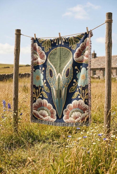 A midnight navy woven cotton blanket featuring a large central raven skull surrounded by rust and ochre flowers, and sage green ferns. Crescent moons are located in the four corners. The blanket has a colorful fringed edge and a textured, loomed appearance. Displayed as a premium gothic botanical home decor piece by Grimoire and Goblet.