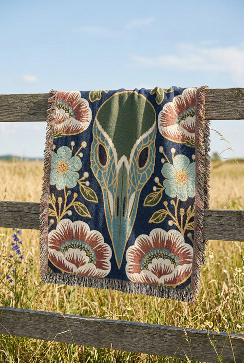 A midnight navy woven cotton blanket featuring a large central raven skull surrounded by rust and ochre flowers, and sage green ferns. Crescent moons are located in the four corners. The blanket has a colorful fringed edge and a textured, loomed appearance. Displayed as a premium gothic botanical home decor piece by Grimoire and Goblet.