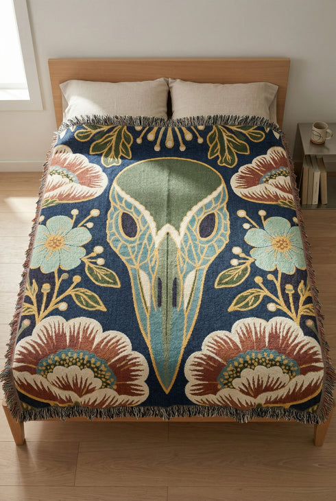 A midnight navy woven cotton blanket featuring a large central raven skull surrounded by rust and ochre flowers, and sage green ferns. Crescent moons are located in the four corners. The blanket has a colorful fringed edge and a textured, loomed appearance. Displayed as a premium gothic botanical home decor piece by Grimoire and Goblet.