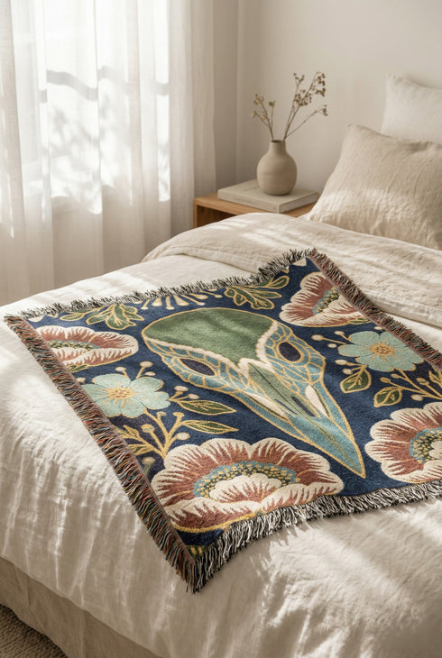 A midnight navy woven cotton blanket featuring a large central raven skull surrounded by rust and ochre flowers, and sage green ferns. Crescent moons are located in the four corners. The blanket has a colorful fringed edge and a textured, loomed appearance. Displayed as a premium gothic botanical home decor piece by Grimoire and Goblet.