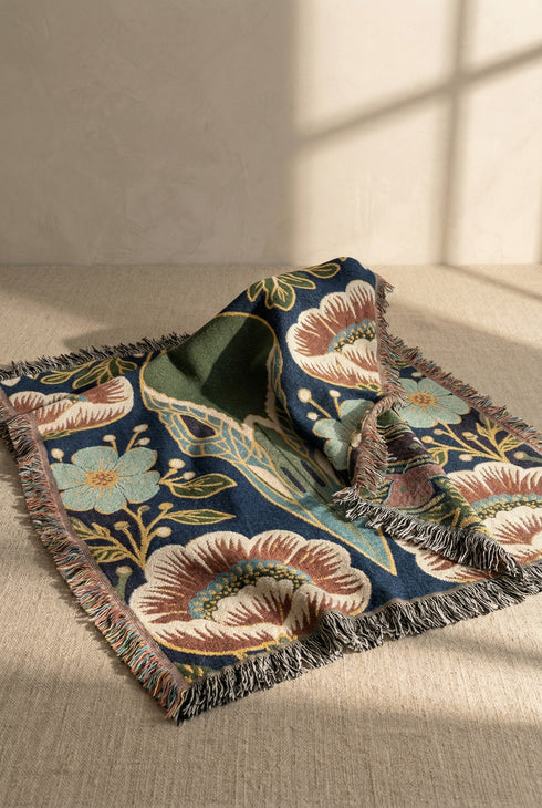 A midnight navy woven cotton blanket featuring a large central raven skull surrounded by rust and ochre flowers, and sage green ferns. Crescent moons are located in the four corners. The blanket has a colorful fringed edge and a textured, loomed appearance. Displayed as a premium gothic botanical home decor piece by Grimoire and Goblet.