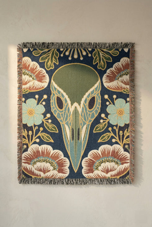 A midnight navy woven cotton blanket featuring a large central raven skull surrounded by rust and ochre flowers, and sage green ferns. Crescent moons are located in the four corners. The blanket has a colorful fringed edge and a textured, loomed appearance. Displayed as a premium gothic botanical home decor piece by Grimoire and Goblet.