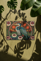 Rectangular low profile doormat printed with a faux embroidery effect that mimics raised crewel stitching. A tall teal blue heron stands at the center among olive green reeds, cattails, and cream water lilies. The rust orange ground is filled with curving botanical vines and small floral medallions, framed by a thin circular border with decorative sunburst corners. A sleek solid black rubber trim finishes all four edges of the one sided print.