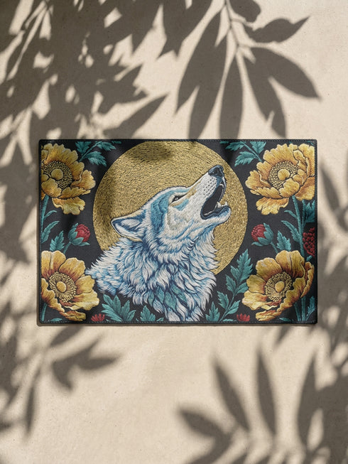 Grimoire and Goblet rectangular doormat featuring a faux embroidery style printed design of a howling wolf with blue and ivory fur, centered in front of a textured antique gold circular moon disc, surrounded by large golden yellow flowers, teal leaves, and small red berry accents on a dark charcoal background. Entryway rug with black trim, non slip backing, and botanical nature inspired mystical design.