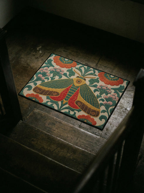 Rectangular low profile doormat printed on a cream ground with a faux embroidery effect that imitates raised satin stitch. A large symmetrical moth sits at the center with teal green wings, mustard yellow upper edges, and deep red lower wing markings. Surrounding it are oversized burnt orange and red blooms, winding green foliage, and clusters of small pink flower buds. A solid black trim frames all four edges of the flat, one sided printed surface.