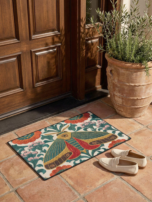Rectangular low profile doormat printed on a cream ground with a faux embroidery effect that imitates raised satin stitch. A large symmetrical moth sits at the center with teal green wings, mustard yellow upper edges, and deep red lower wing markings. Surrounding it are oversized burnt orange and red blooms, winding green foliage, and clusters of small pink flower buds. A solid black trim frames all four edges of the flat, one sided printed surface.