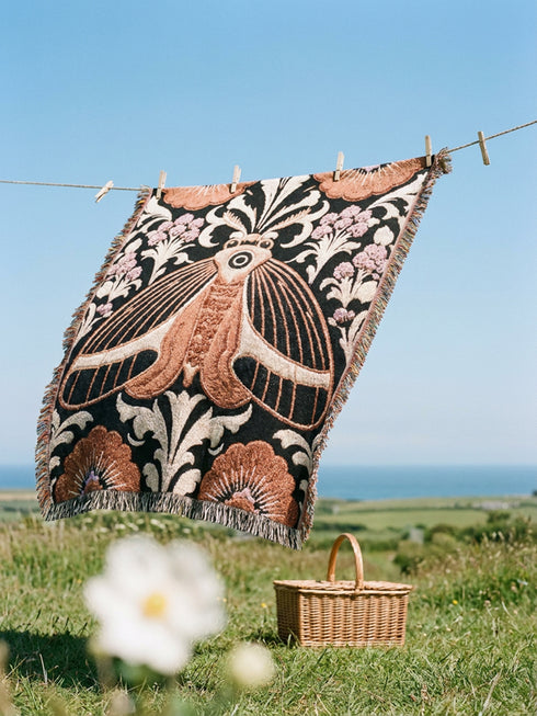 Woven cotton throw blanket with fringed edges and a black ground. At the center is a large moth with copper, brown, and cream wings marked by fine stripes and a concentric eyespot, with feathered antennae. Cream acanthus scrollwork fans out around it in the Art Nouveau manner, joined by clusters of lilac and mauve flowers and copper-brown blossoms with pale rays at the top and bottom. The pattern is jacquard woven, and the reverse shows a mirrored, softer version of the design.