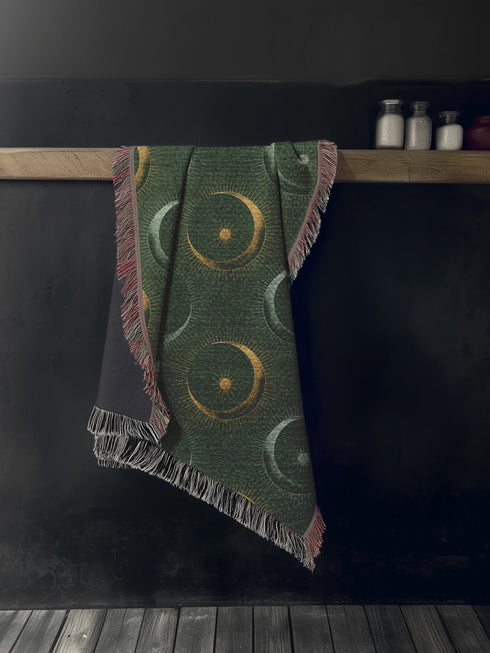 Forest green cotton throw blanket with woven silver moon phases and golden sun motifs, finished with multicolored fringe trim.