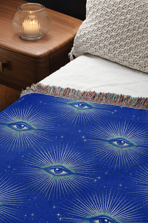 Woven cotton throw blanket with a deep blue background featuring a repeated pattern of all-seeing eyes in gold and seafoam green. The blanket has a colorful fringed edge.