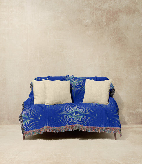 Woven cotton throw blanket with a deep blue background featuring a repeated pattern of all-seeing eyes in gold and seafoam green. The blanket has a colorful fringed edge.