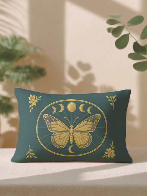 A rectangular lumbar pillow cover, measuring 20 by 12 inches, is displayed against a neutral background. The pillow cover is made of a deep teal green fabric with a subtle, linen-like texture. In the center, a large, intricately detailed butterfly with spread wings is printed in a shimmering gold color. The butterfly is framed by a golden oval which also contains a series of gold moon phase symbols, showing the full lunar cycle. The four corners of the pillow is adorned with gold floral design.