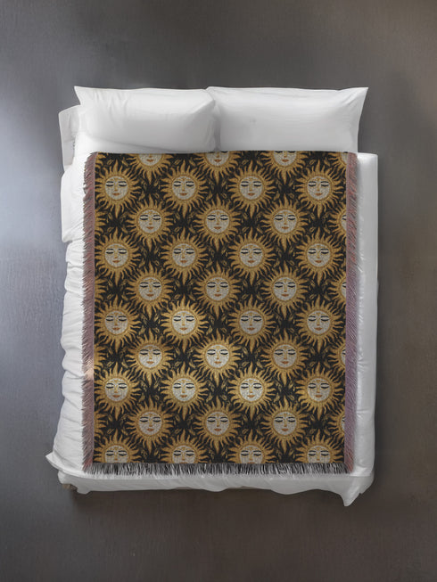 A woven cotton throw featuring a celestial sun face pattern in gold and black tones, framed by soft fringe.