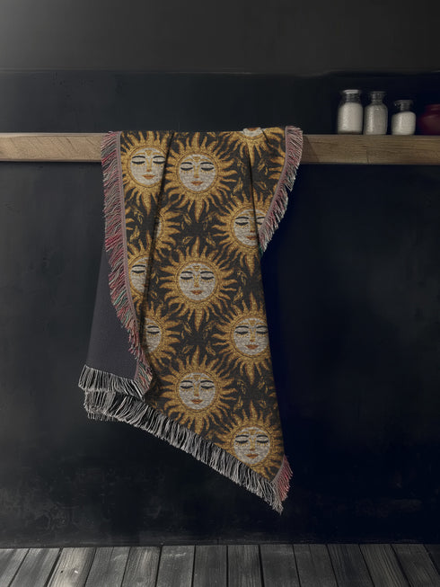A woven cotton throw featuring a celestial sun face pattern in gold and black tones, framed by soft fringe.