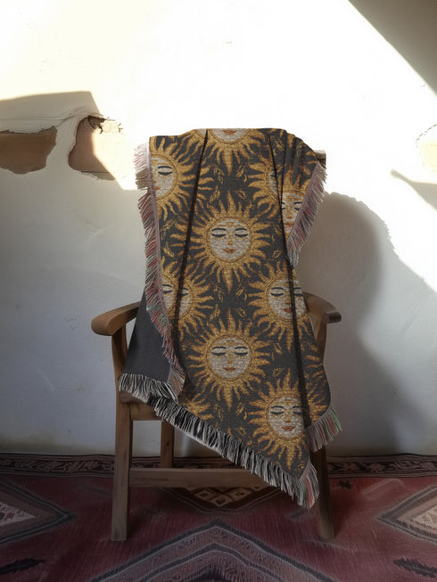 A woven cotton throw featuring a celestial sun face pattern in gold and black tones, framed by soft fringe.