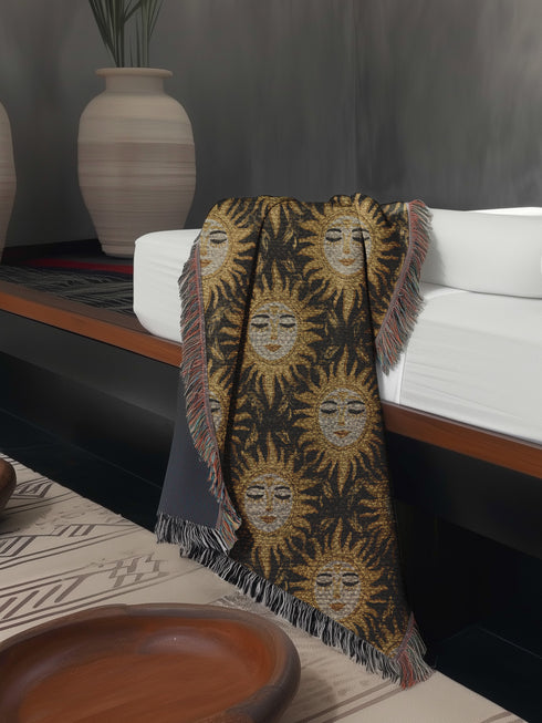 A woven cotton throw featuring a celestial sun face pattern in gold and black tones, framed by soft fringe.