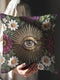 Grimoire and Goblet gothic pillow cover featuring a central evil eye surrounded by radiating gold sunburst rays, framed by white daisy flowers, magenta floral blooms, and green botanical foliage on a black background, designed with a highly detailed faux embroidery print that mimics stitched texture while remaining smooth, arranged in a symmetrical medallion composition, creating a dark academia and witchy botanical aesthetic decorative cushion cover